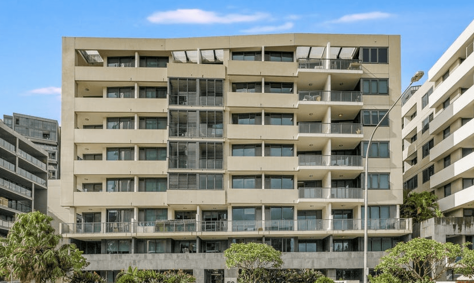 Front view of a modern multi-story apartment building with balconies, large windows, and surrounding greenery.